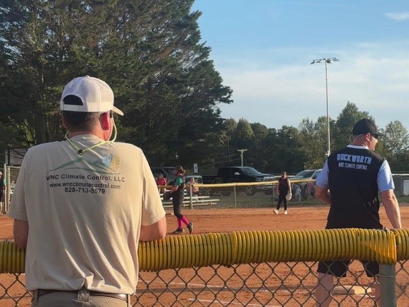 WNC Climate Control team member in a company shirt watching a youth softball game from the fence line, with a coach in a WNC Climate Control jersey on the field.